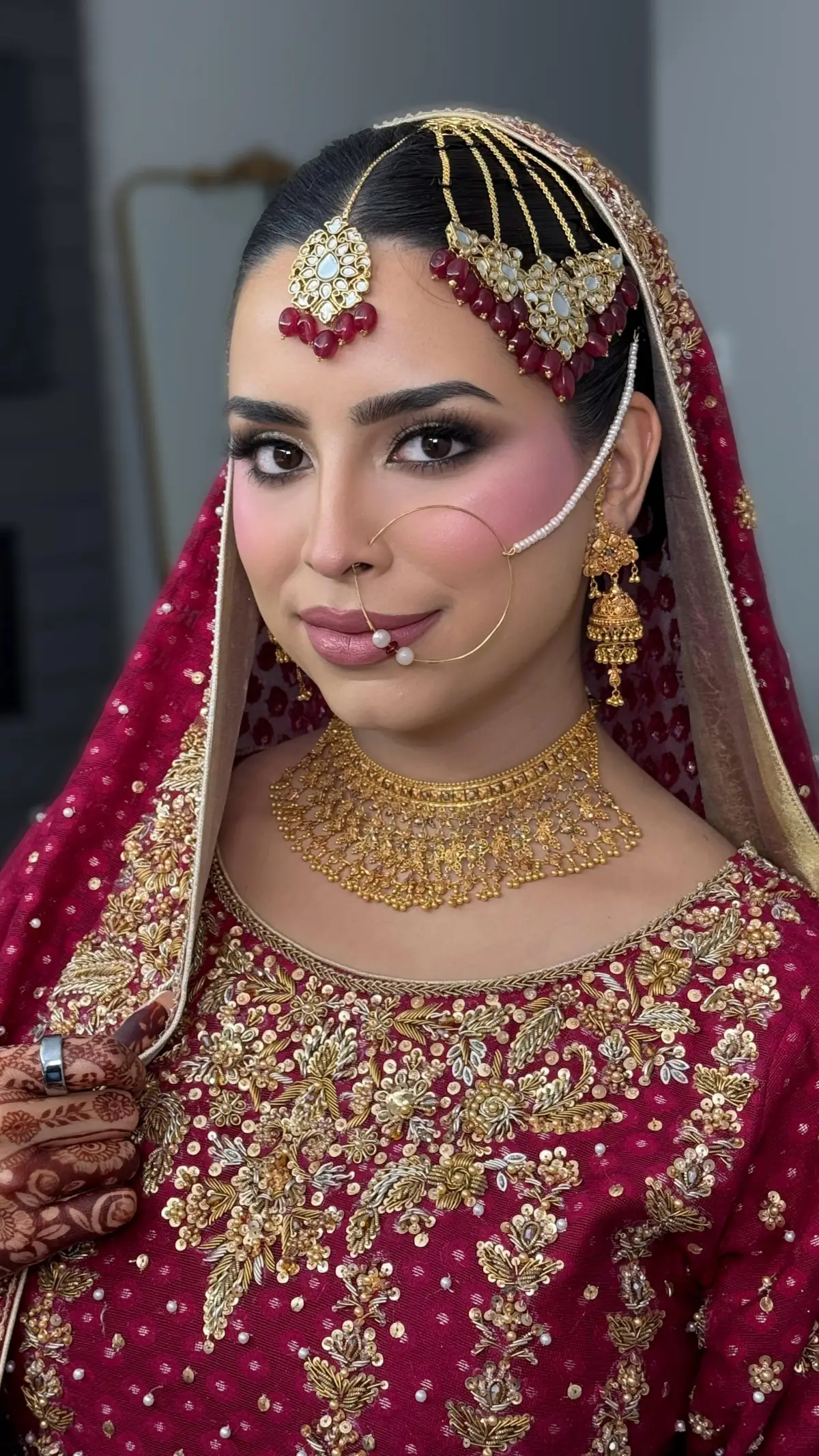 Bride in crimson and gold with a matha patti and nose chain, mehndi hands resting in her lap.
