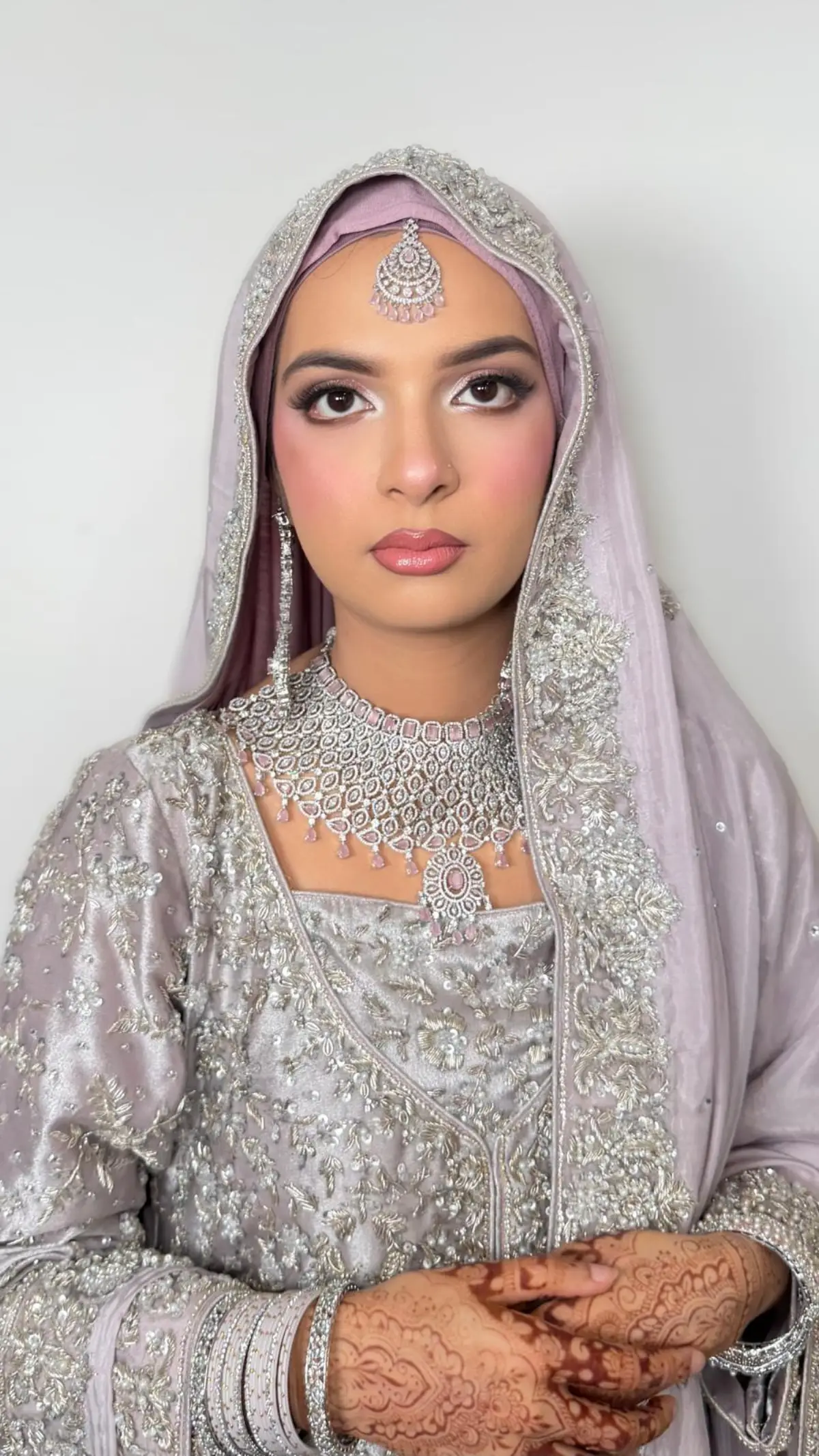 Hijabi bride in lilac and silver, hijab draped and pinned into a smooth crown, henna on her hands.