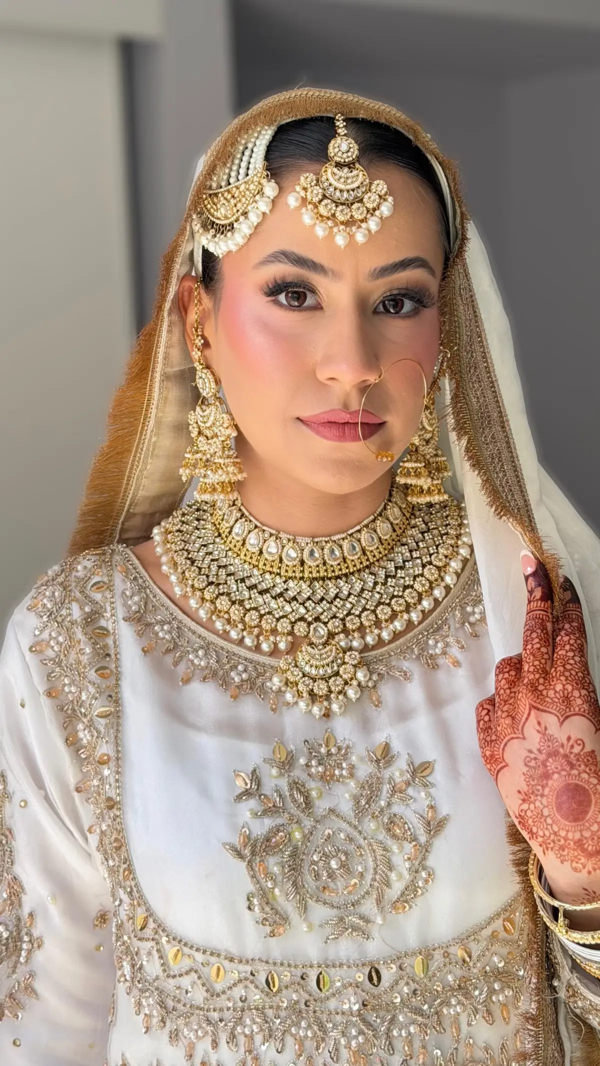 Bride in ivory and gold with a layered kundan necklace, fresh mehndi on her hands and a white dupatta over her hair.