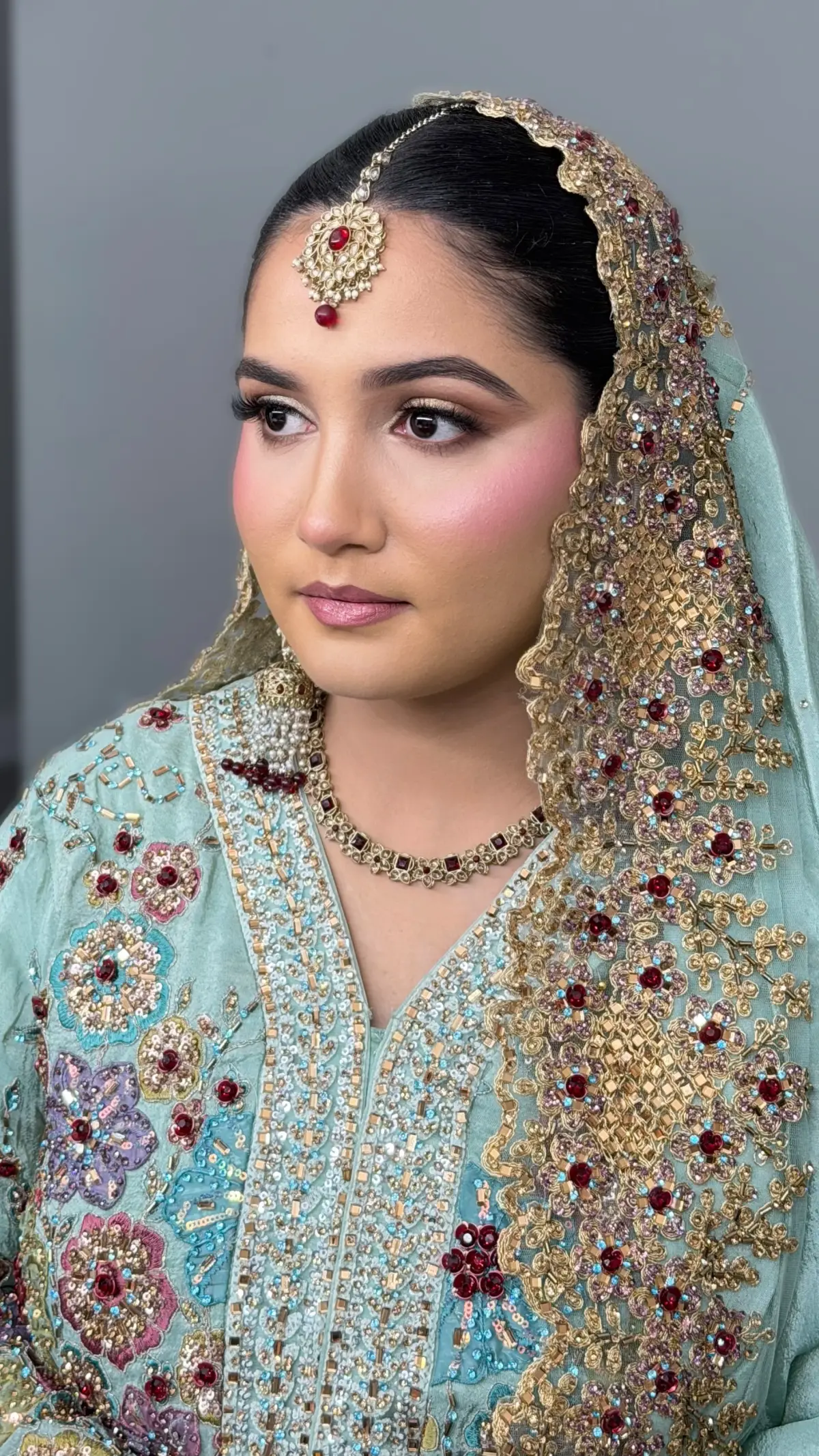 Bride in mint and gold with an embroidered dupatta draped over her head, maang tikka and soft rose makeup.