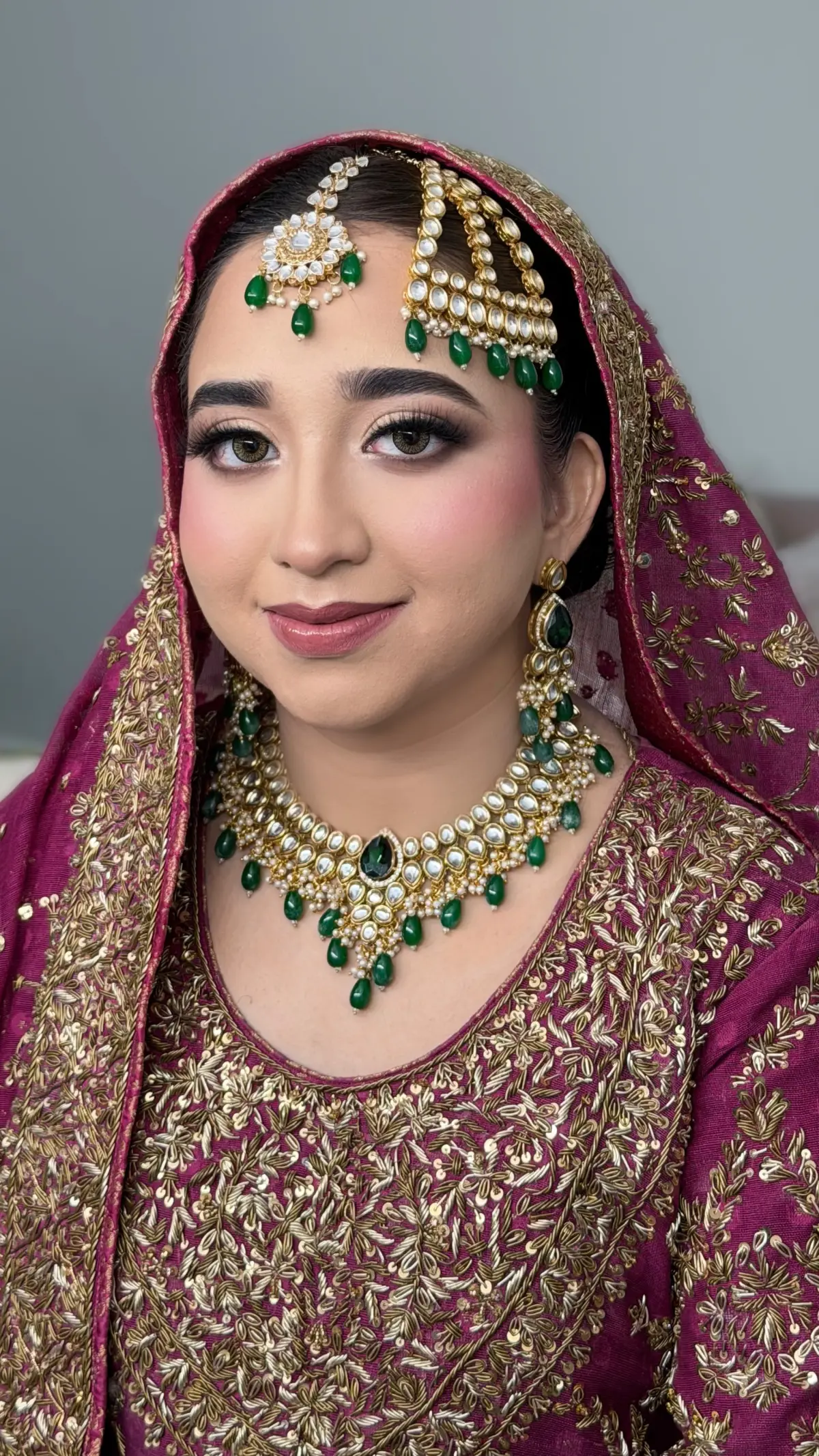 Bride in deep plum and gold with emerald green kundan jewellery and a dupatta pinned over her hair.