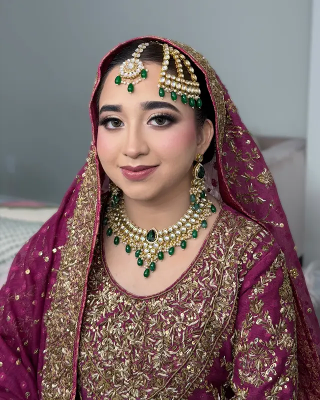 Bride in deep plum and gold with emerald green kundan jewellery and a dupatta pinned over her hair.
